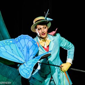 An actor in a blue costume and yellow hat holding a large butterfly fixed on a rod