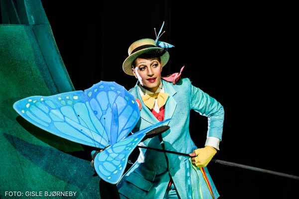 An actor in a blue costume and yellow hat holding a large butterfly fixed on a rod