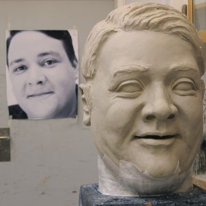 A big head is being modelled and casted. There is a enlarged photograph of the actor in the background